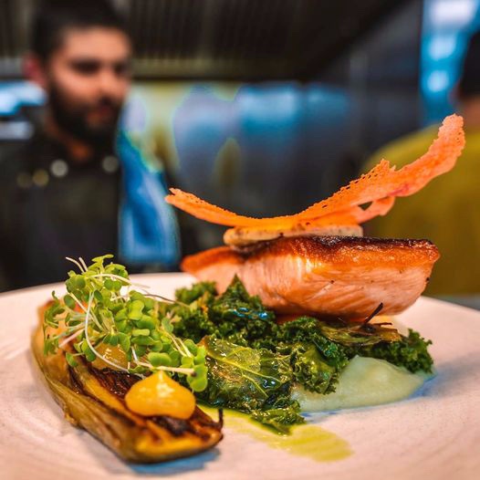 Akaroa Salmon with parsnip puree, braised kale, beetroot coral, and caper butter on a plate with chef in the background.